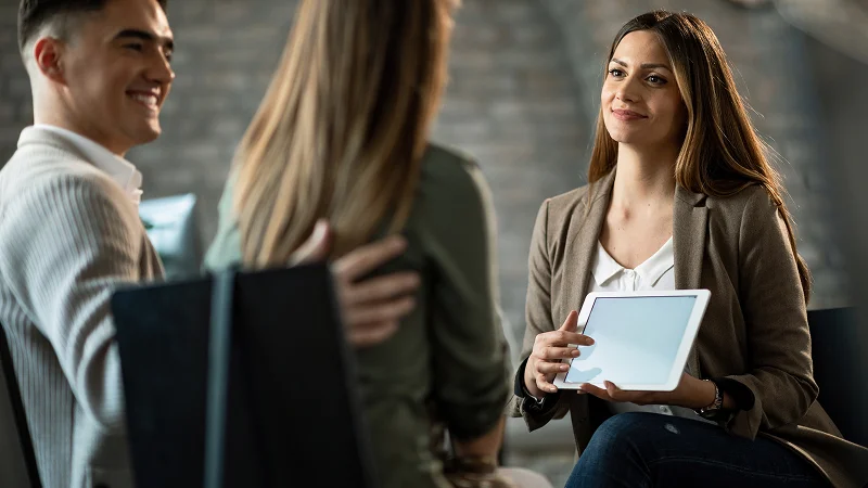 A female professional in a blazer presenting information on a tablet to a couple.