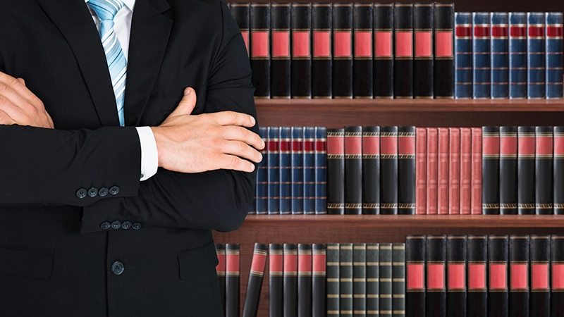 Close-up Of Male Lawyer With Arm Crossed Standing In Front Of Book Shelf.